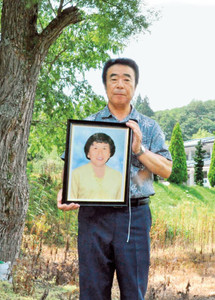 渡辺はま子さんが亡くなった場所付近で、遺影を持つ夫の幹夫さん＝７月、福島県川俣町で